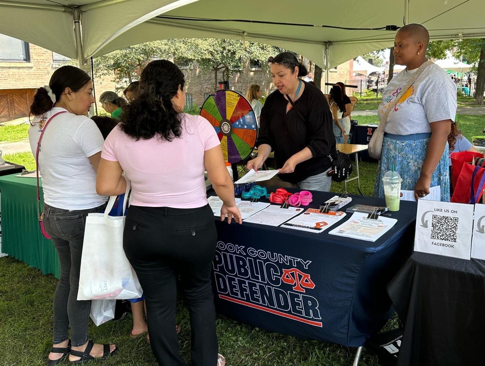 Two young women speak to representatives from the Public Defender's office at an outdoor fair in Chicago called Fiesta del Sol. Two women representing the Public Defender's Office are providing resources from a table under a tent.