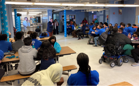 Students sit at long tables in a cafeteria, listening to a speaker from the Public Defender's Office who stands in the center of the room.